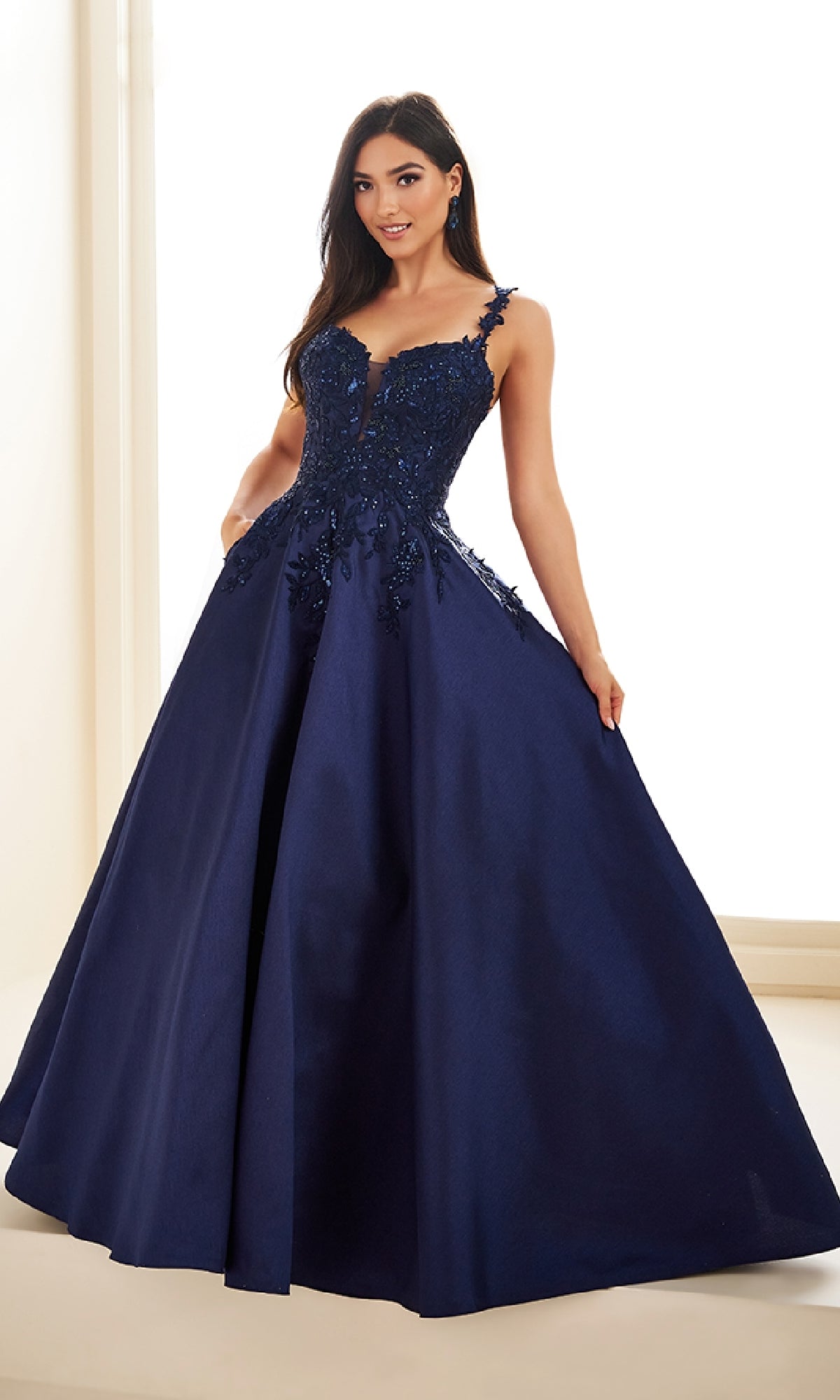 A woman wearing the Ellie Wilde EW36094 long prom dress in dark blue with lace embellishments and a corset bodice stands indoors against a light background.